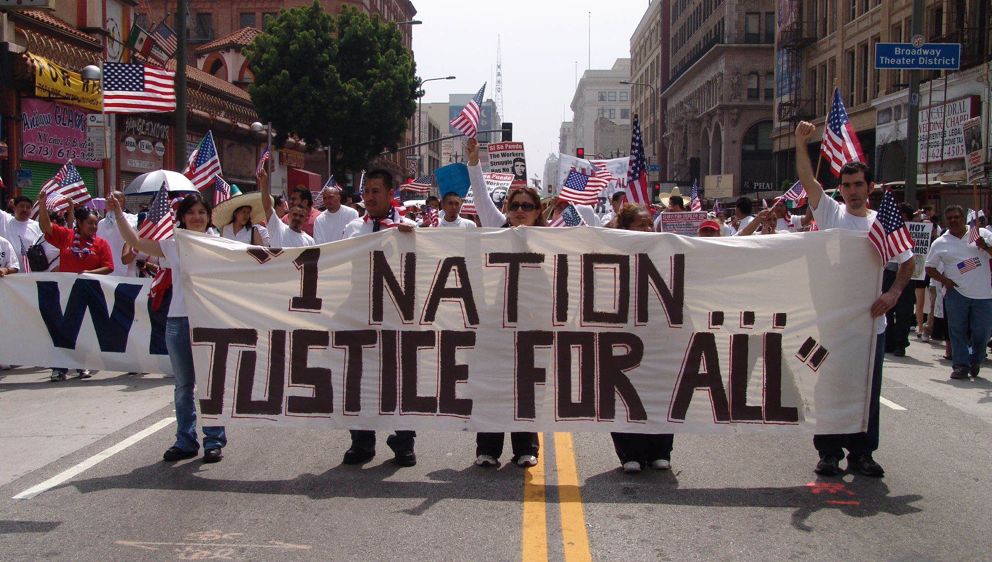 a large crowd marches in a downtown area holding a banner that says '1 Nation...Justice for All'