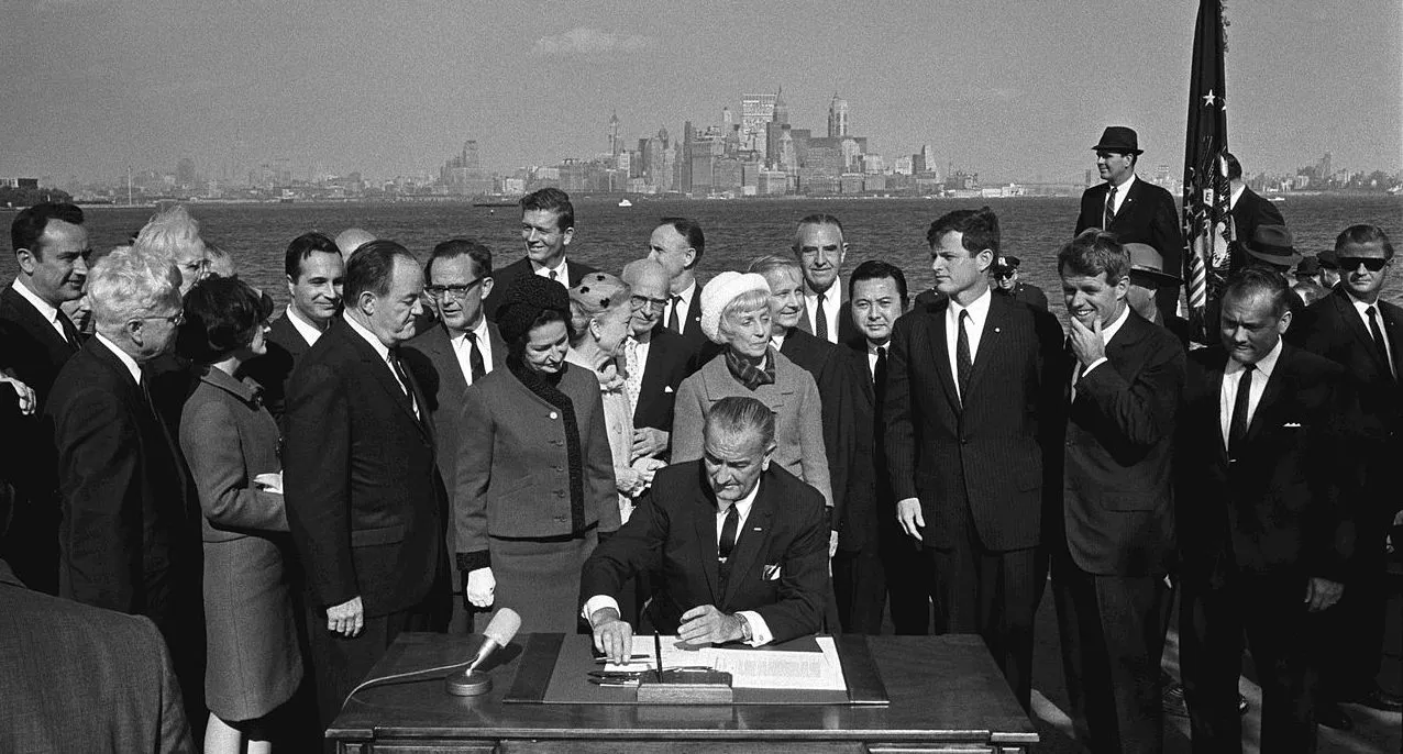 President Johnson signs the 1965 Immigration Act surrounded by dignitaries outside at Ellis Island, NY.